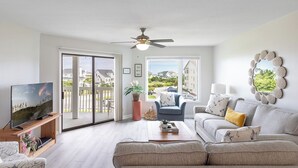 A modern living room with a television, sofa, and a view of a residential area through large windows. - Point Emerald E-202, Oceanside Condo in Emerald Isle, NC! (Emerald Isle)