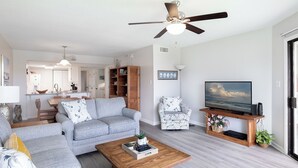 A modern living room with a grey sofa, a wooden coffee table, a TV on a wooden stand, and a ceiling fan. - Point Emerald E-202, Oceanside Condo in Emerald Isle, NC! (Emerald Isle)