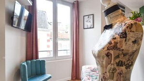 A room with a blue chair, a bed with a floral bedspread, a television mounted on the wall, and a window with curtains.
