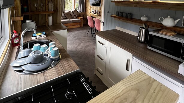 A compact kitchen with a stove, sink, and wooden countertops. There are shelves with kitchenware and a seating area visible in the background.