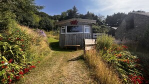 A small wooden cabin with a window and a bench in front, surrounded by a garden with various plants and flowers.