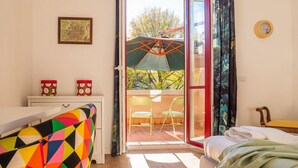 A room with a bed, a colorful chair, a small table with decorative items, a framed picture on the wall, and a sliding glass door leading to an outdoor area with a chair and umbrella. - The Best Rent - Apartment close to Porta Romana (Milano)