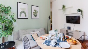 Uma sala de estar moderna com uma mesa de jantar redonda posta para o café da manhã, um sofá com almofadas, uma TV montada na parede e um vaso de planta.