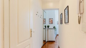 Um corredor com piso de madeira, paredes brancas e obras de arte emolduradas. Há uma porta à esquerda e um radiador com plantas à direita.