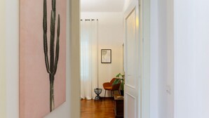 Um corredor com piso de madeira, uma obra de arte de cactos à esquerda, uma cadeira, uma mesinha e um quadro na parede.