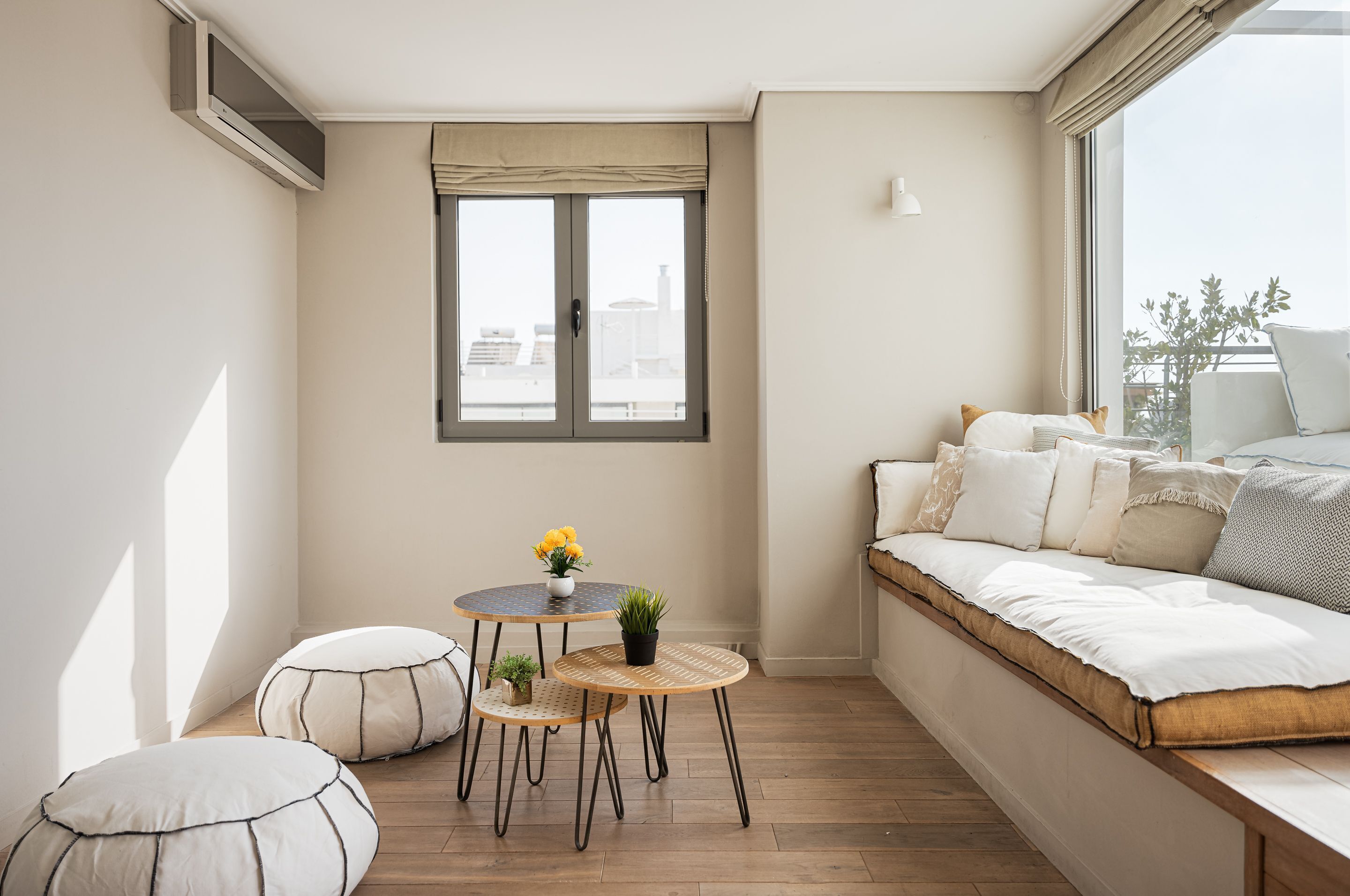 A small, cozy room with a built-in bench, two round tables, and a window with a view of buildings.