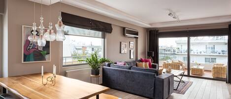 Um espaço de estar moderno com uma mesa de jantar de madeira, sofás cinzentos e uma grande janela com vista para a cidade.