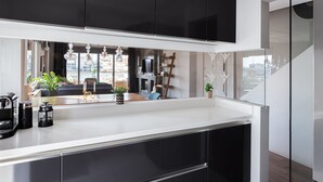 A modern kitchen with dark cabinets, a white countertop, and a view of a living area with a sofa and plants. - Glyfada Luxury Living (Glyfada)
