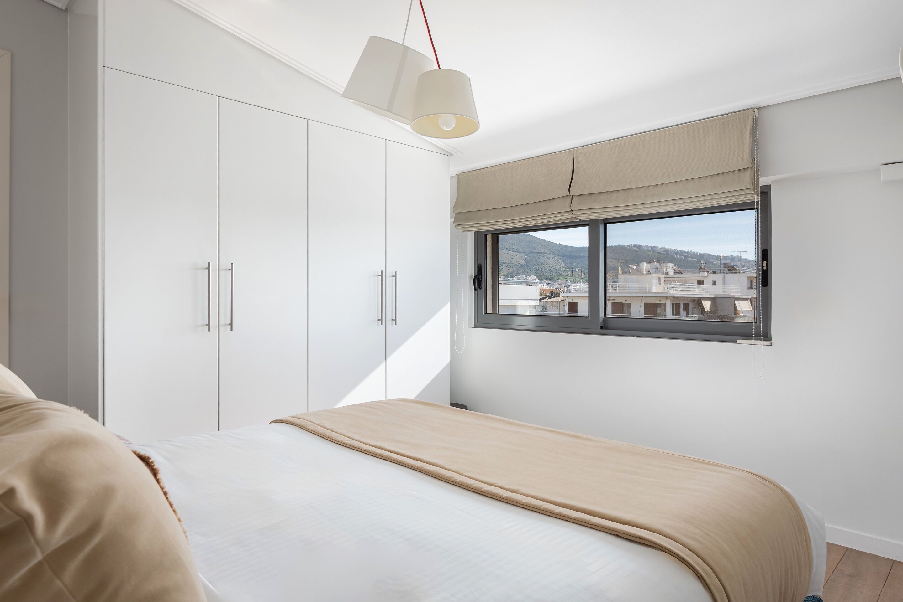 Une chambre à coucher avec un lit, une armoire blanche et une fenêtre donnant sur des immeubles et des montagnes.