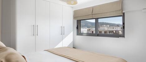 Une chambre à coucher avec un lit, une armoire blanche et une fenêtre donnant sur des immeubles et des montagnes.