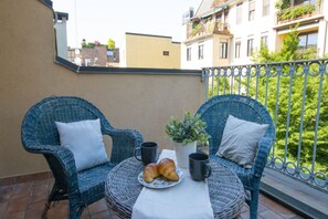 Een balkon met rieten stoelen, een klein tafeltje, een bloempot en croissants.