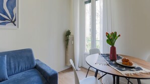 A living room with a blue sofa, a white table with a red vase and tulips, and a window with curtains. - The Best Rent - Apartment in Piazza Cinque Giornate (Milano)