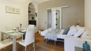 Een moderne, goed verlichte kamer met een eettafel, witte stoelen, een bank en een bed.