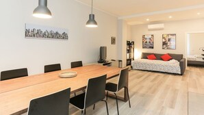 Un salon moderne avec une table à manger en bois, des chaises noires, un canapé gris agrémenté de coussins rouges et une œuvre d’art représentant un paysage urbain accrochée au mur.