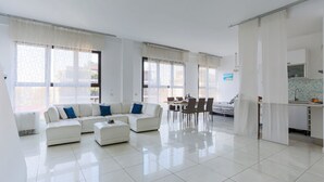 Een moderne woonkamer met een witte zithoek, een eethoek met een tafel en stoelen, en een keuken met een spoelbak en kastjes.