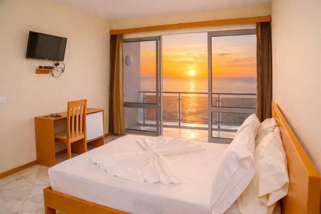 Een hotelkamer met een bed, een bureau, een stoel, een televisie en een balkon met uitzicht op een zonsondergang.