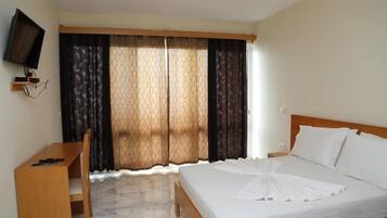 A hotel room with a bed, a desk, a chair, a TV mounted on the wall, and a window with patterned curtains.