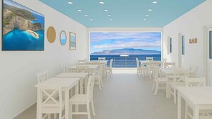 A dining area with white tables and chairs, a large window with a view of the sea and mountains, and a painting of a coastal scene. - Cruzeiro Guest House (São Filipe)