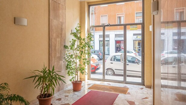 Entrada de hotel con suelo de baldosas, plantas en macetas y vista de coches aparcados afuera.