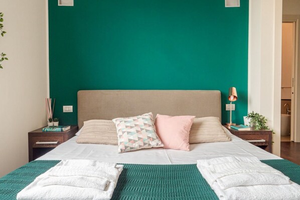 A neatly made bed with two pillows, a teal wall, and a small bedside table with a lamp.