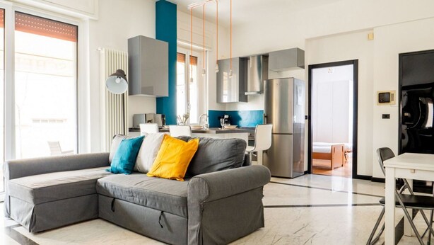 A modern living room with a grey sectional sofa, a dining table, and a kitchen area with stainless steel appliances.
