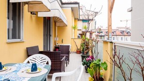 A balcony with a table set for two, wicker chairs, potted plants, and a view of the cityscape.