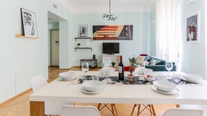 Un espace de salle à manger moderne, avec une table blanche dressée pour quatre personnes, un téléviseur au-dessus et des œuvres d’art accrochées aux murs.