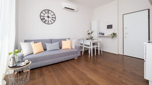 A modern living room with a grey sofa, a round wall clock, a dining table with chairs, and a white door. - The Best Rent - Bright one-bedroom apartment in Milan (Milano)
