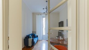 A modern living room with a wooden herringbone floor, a blue sofa with cushions, a dining table with a red chair, and a kitchen area visible through an open door. - The Best Rent - Flat with 1 bedroom in Moscova area (Milano)