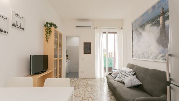 A modern living room with a grey sofa, a flat-screen TV, a white dining table, and a kitchen area in the background. - The Best Rent - Two bedrooms apartment near metro station Maciachini (Milano)