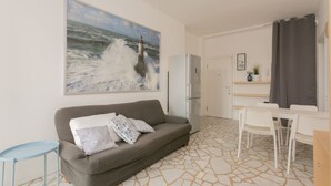 A modern living room with a grey sofa, a small blue side table, a white dining table with chairs, a refrigerator, and a large painting of a lighthouse on the wall. - The Best Rent - Two bedrooms apartment near metro station Maciachini (Milano)