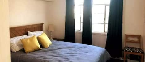 A bedroom with a bed, a chair, a window with curtains, and an air conditioning unit.