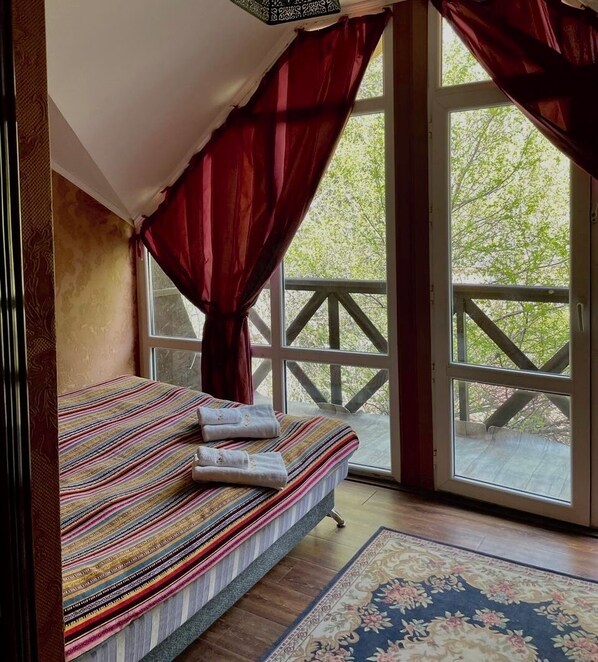 A room with a bed, a window with curtains, and a view of greenery outside.
