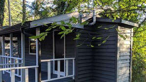 Une petite cabane de couleur sombre, dotée d’une rambarde blanche et d’une porte, entourée d’arbres.