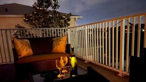 Een balkon met een rieten bank, twee kussens en een glazen tafel met wijnglazen.