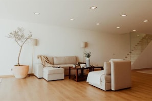 Uma sala de estar moderna com um sofá, poltrona e mesa de centro, uma planta em vaso e uma escada ao fundo.