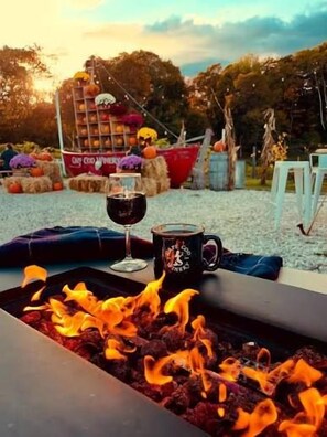 A fire pit with a glass of red wine and a mug, surrounded by outdoor seating and hay bales.