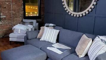 A modern living room with a grey sofa, a round mirror with a bead trim, and a brick wall.