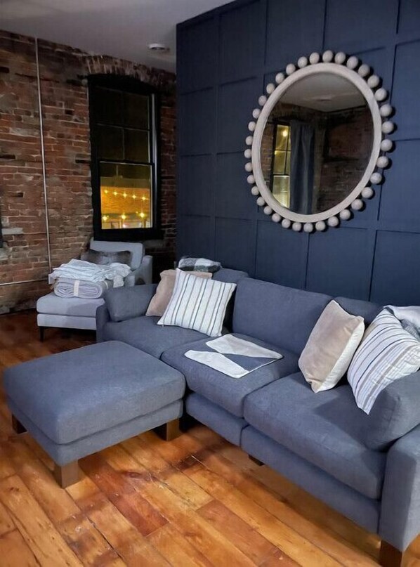 A modern living room with a grey sofa, a round mirror with a bead trim, and a brick wall. - Mill Street Rentals #2 (North Utica)