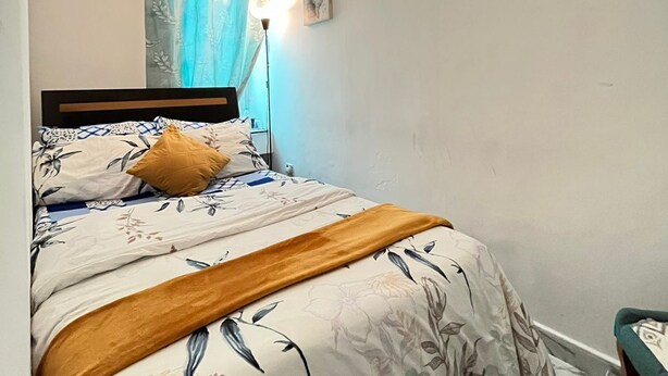 Un dormitorio con cama, mesita de noche, lámpara, ventana con cortinas e imagen colgada en la pared.