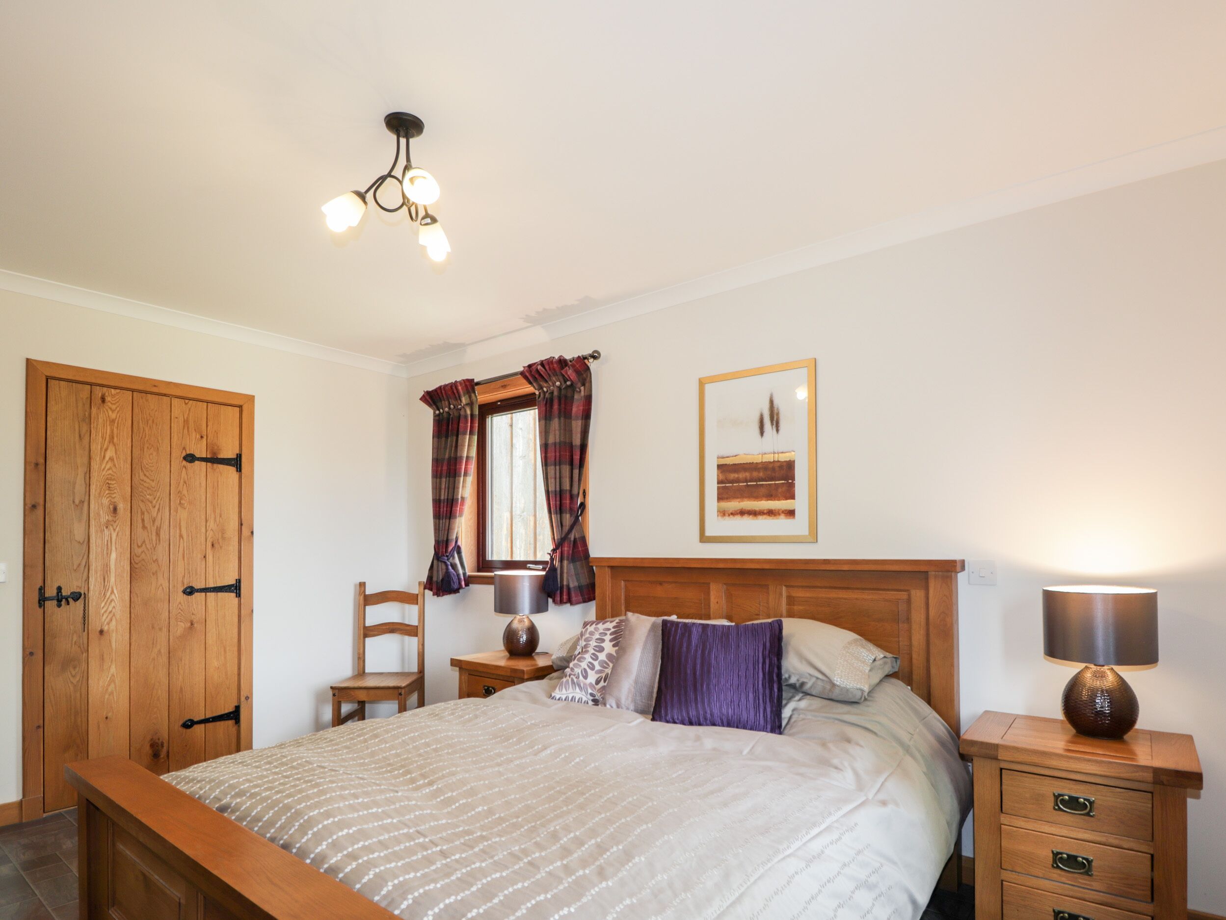 A bedroom with a wooden bed, a nightstand with a lamp, a chair, a window with curtains, and a framed picture on the wall.