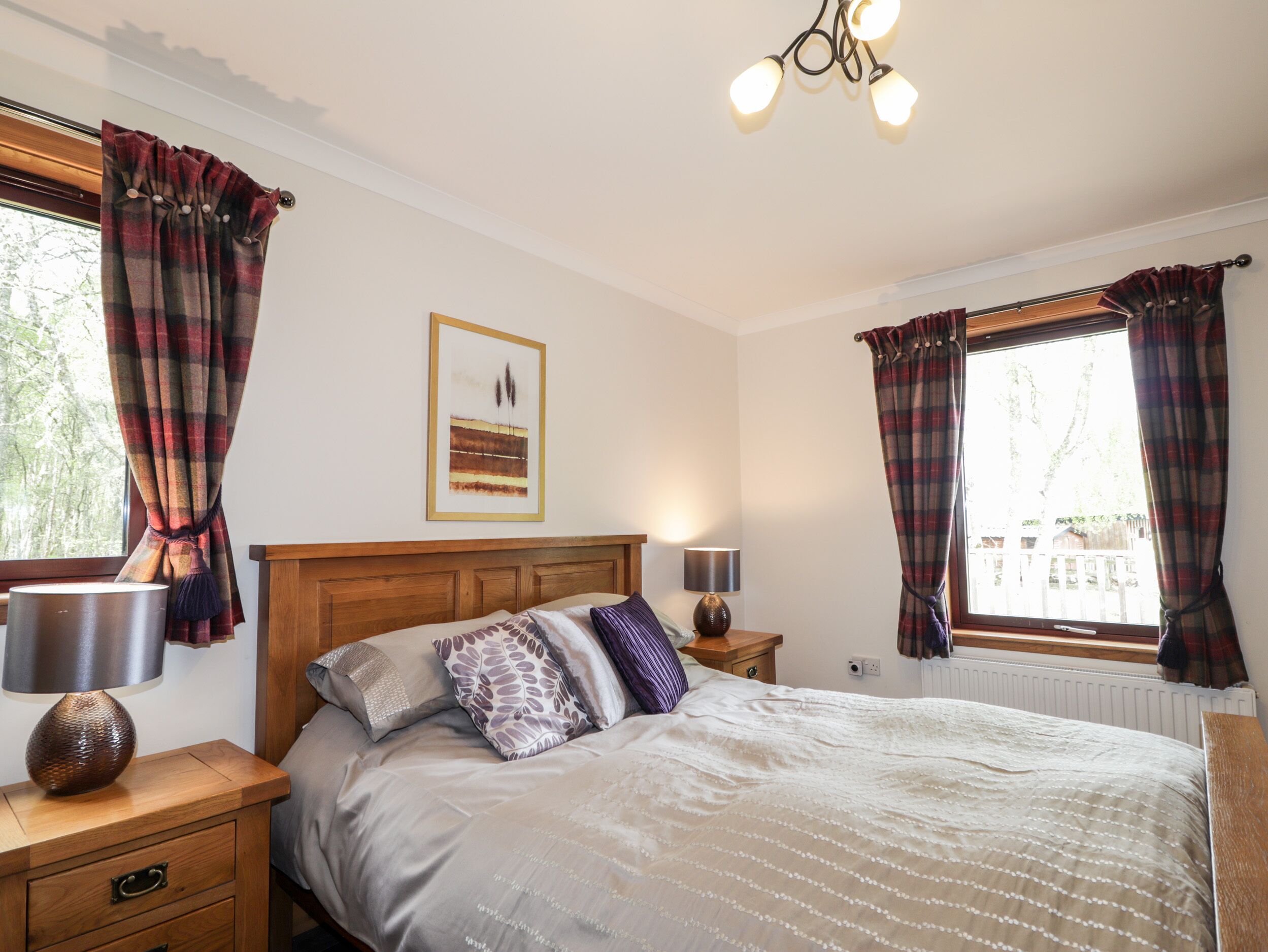 A bedroom with a bed, two bedside tables with lamps, a wooden headboard, a framed picture on the wall, and plaid curtains on the windows.