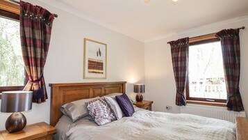A bedroom with a bed, two bedside tables with lamps, a wooden headboard, a framed picture on the wall, and plaid curtains on the windows.