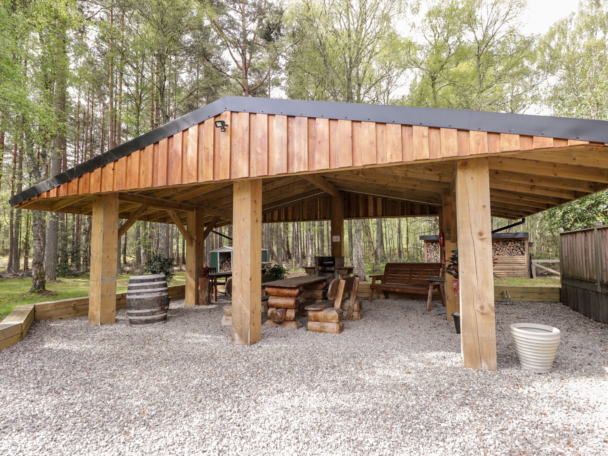 A covered outdoor seating area with wooden benches and tables, surrounded by gravel and trees.