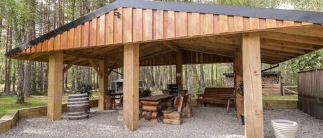 A covered outdoor seating area with wooden benches and tables, surrounded by gravel and trees.