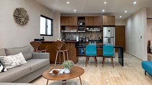 A modern living room with a sofa, coffee table, and two chairs. A kitchen with wooden cabinets and appliances is visible in the background. - Newly build 2 bedroom, luxury apartment a few steps from beach. (Puerto Morelos)