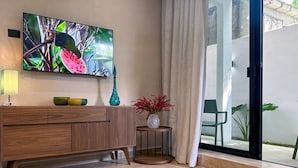 A modern living room with a wooden cabinet, a flat-screen TV, a small side table with a vase, and a view of the outdoors through a glass door. - Newly build 2 bedroom, luxury apartment a few steps from beach. (Puerto Morelos)
