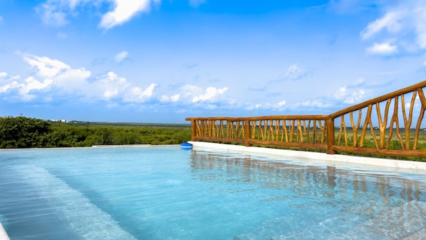 A clear blue infinity pool with a wooden deck railing, surrounded by lush greenery and a clear sky. - Newly build 2 bedroom, luxury apartment a few steps from beach. (Puerto Morelos)