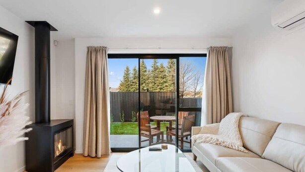 Un salon moderne avec une table basse en verre, un canapé beige, une cheminée et une vue sur un jardin avec une terrasse en bois et des chaises.