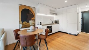 Une cuisine moderne avec des meubles blancs, un coin repas doté d’une table ronde et de chaises marron, et un grand tableau abstrait accroché au mur.
